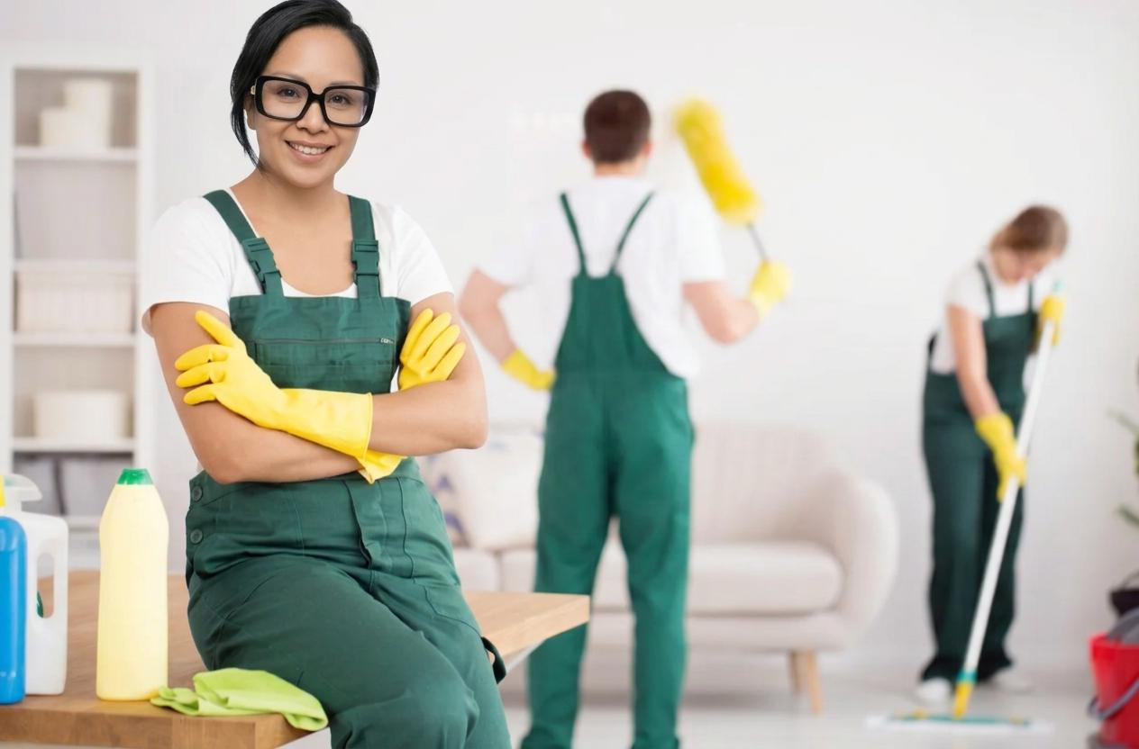 Carla in green overalls and yellow gloves, smiling, while her team cleans the living room behind her