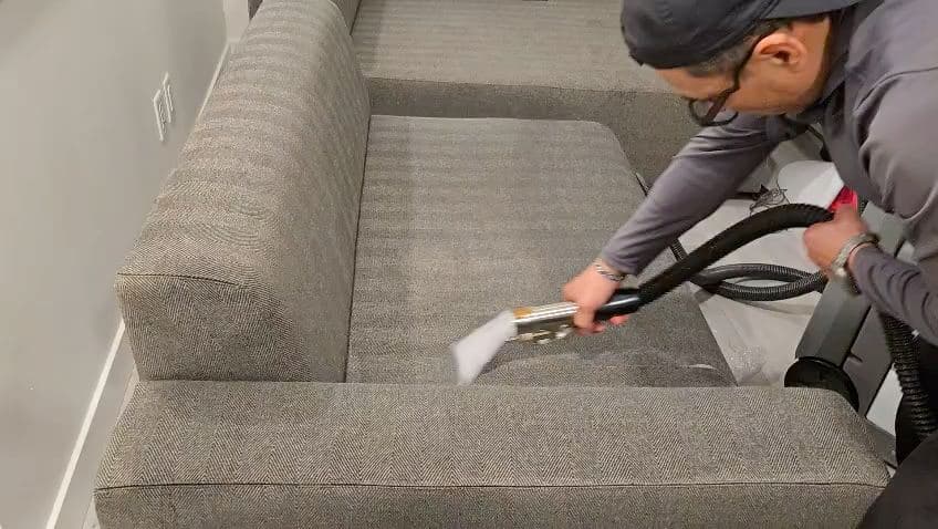 Cleaner running a hot-water extraction wand along a grey sofa cushion